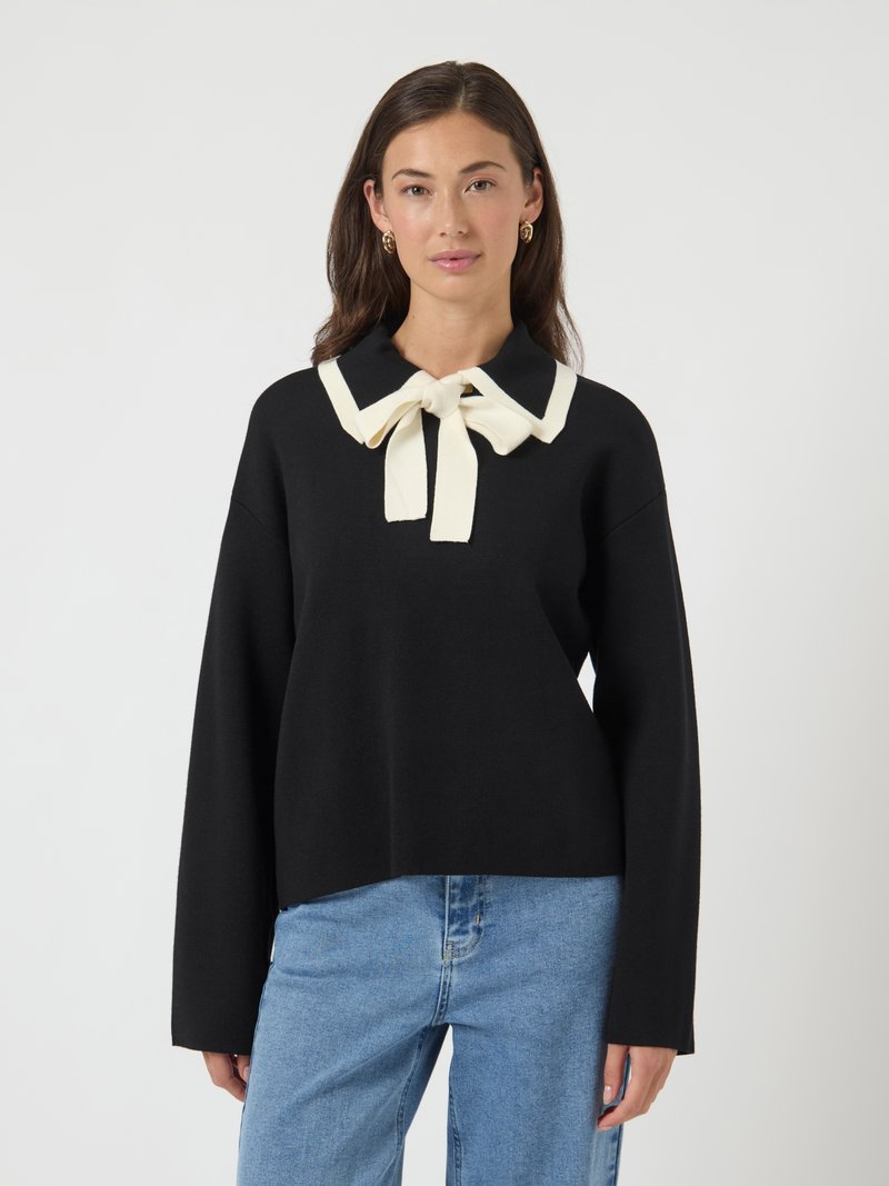 Kvinne iført svart langermet topp med hvit krage og sløyfedetalj, sammen med lyseblå jeans, stående mot hvit bakgrunn.