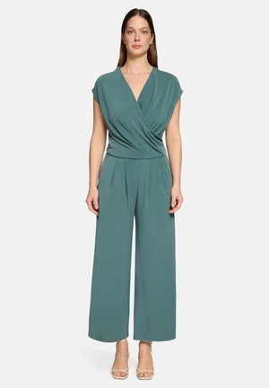 Frau mit langen, glatten Haaren, die einen tealfarbigen ärmellosen Wickel-Top-Jumpsuit und beige offene High Heels trägt, stehend vor einem einfarbigen hellen Hintergrund.