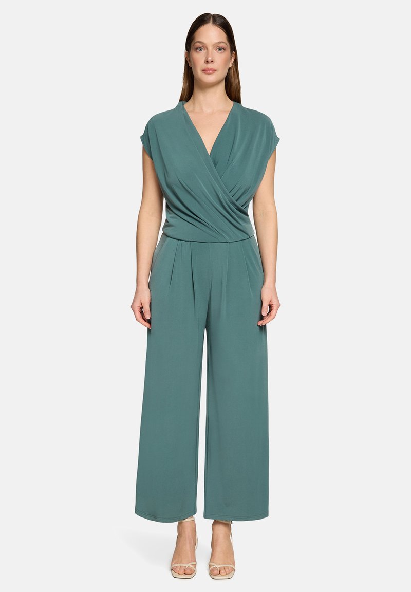 Femme aux longs cheveux lisses portant une combinaison-pantalon sans manches de couleur sarcelle avec un haut cache-cœur et des talons ouverts beiges, debout devant un fond clair uni.
