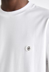 Tricou alb din bumbac, cu guler rotund și un mic logo circular în nuanțe de argintiu-gri, brodat pe material.