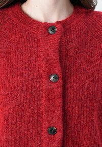 Cardigan en tricot rouge avec un motif texturé, encolure ronde et boutons noirs sur le devant. Tissu en laine douce.