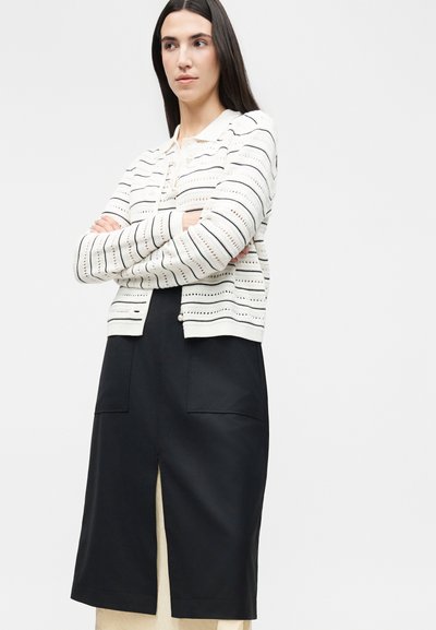 Γυναίκα με μακριά σκούρα μαλλιά που φοράει λευκή ριγέ πλεκτή ζακέτα και μαύρη μίντι φούστα με μπροστινές τσέπες και σκίσιμο εμπρός, με σταυρωμένα τα χέρια.