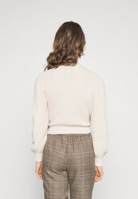 Krémový pletený svetr s vysokým límcem a balónovými rukávy, doplněný hnědými kalhotami se vzorem káro a elastickým pasem. Texturovaný, volný střih.