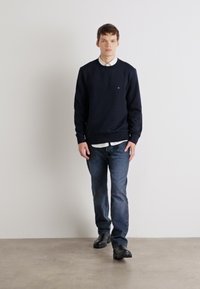 Marineblå genser med rund hals, brukt over en hvit skjorte med krage. Kombinert med blå jeans og svarte sko. Enkelt, uformelt antrekk.