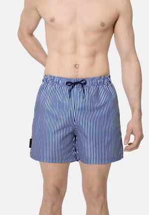 Hombre sin camiseta con pantalones cortos de baño a rayas verticales azules y blancas con cordón negro sobre un fondo liso.