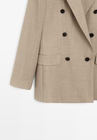 Beige dubbelknäppt kavaj med svarta knappar, långa ärmar och ficklock, visad mot en vit bakgrund.