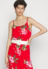 Vestido floral vermelho com alças finas, apresentando padrões florais em rosa e verde. Um cinto largo branco aperta na cintura. Textura de tecido suave.