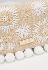 Borsa a tracolla intrecciata beige con accenti floreali bianchi e marroni, una placca con logo ALDO dorata e fascetta in pompon bianca lungo il bordo inferiore.