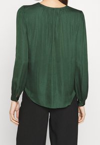 Blusa lunga verde scuro a maniche lunghe realizzata in tessuto morbido, con scollatura rotonda e dettagli arricciati sulle spalle e sulla parte posteriore.