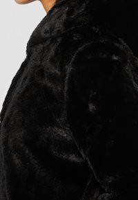 Svart faux fur-kappa med en mjuk, texturerad yta, klassisk krage och en strukturerad passform. Synliga sömmar framhäver den skräddarsydda designen.