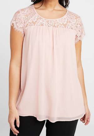 Vrouw draagt een lichtroze blouse met bloemige kanten korte mouwen en een inzetstuk, gecombineerd met een zwarte broek.