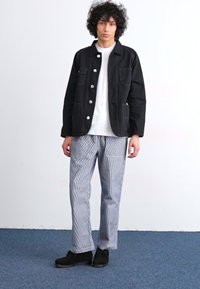 Black denim jacket with white buttons, paired with striped blue and white trousers. Black shoes complete the outfit. Simple and modern design.