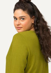 Junge Frau mit langen lockigen dunklen Haaren in einem grünen Oberteil, die über ihre Schulter blickt, vor einem schlichten hellen Hintergrund.