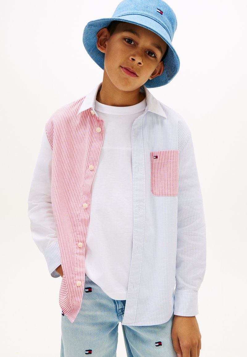 Αγόρι με μπλε καπέλο τύπου κάπα, λευκό πουκάμισο με κόκκινο και λευκό ριγέ πουκάμισο πάνω από αυτό, και ανοιχτό μπλε τζιν με κεντημένα λογότυπα.