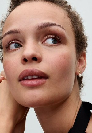 Nahaufnahme eines Gesichts mit heller Haut, hellbraunen Locken, blauen Augen, das einen kleinen goldenen Ohrring mit einem Perlenakzent und minimales Make-up trägt.