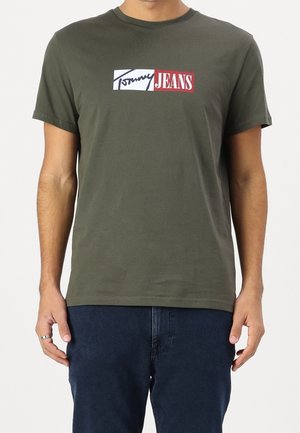 Oliwkowa bawełniana koszulka z krótkim rękawem z graficznym logo w białym i czerwonym kolorze, na którym widnieje napis "Tommy JEANS" w prostokątnym formacie.