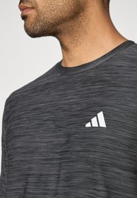 Harmaa urheilupaita, jossa on teksturoitu kuvio ja valkoinen logo vasemmassa rinnassa. Lyhyet hihat ja klassinen pyöreä kaula-aukko.