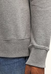 Sweat-shirt gris en tissu doux et texturé avec des poignets et un ourlet côtes. Présente une coupe décontractée, avec un gros plan sur le détail de la manche et du bas.
