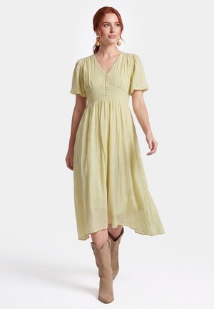 Frau mit roten Haaren, die ein hellgelbes Midikleid mit Knöpfen vorne und Puffärmeln trägt, kombiniert mit beigen Cowboy-Stiefeln, schaut zur Seite.