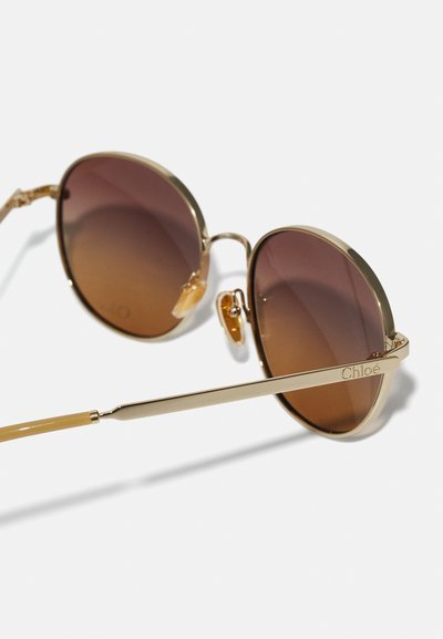 Ochelari de soare avion cu tonuri aurii și lentile cu gradient maro. Dispune de o coroană subțire, texturată, și un accent cu logo pe braț. Include suporturi pentru nas.