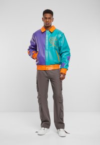 Barevná kožená bomber bunda s fialovými a tyrkysovými panely, oranžovými akcenty a texturovanými pletenými manžetami; doplněná šedými cargo kalhotami a bílými teniskami.