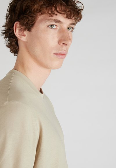 T-shirt beige a collo a V realizzato in un tessuto morbido, con dettagli di cucitura minimal e un design aderente. Modello mostrato di profilo.