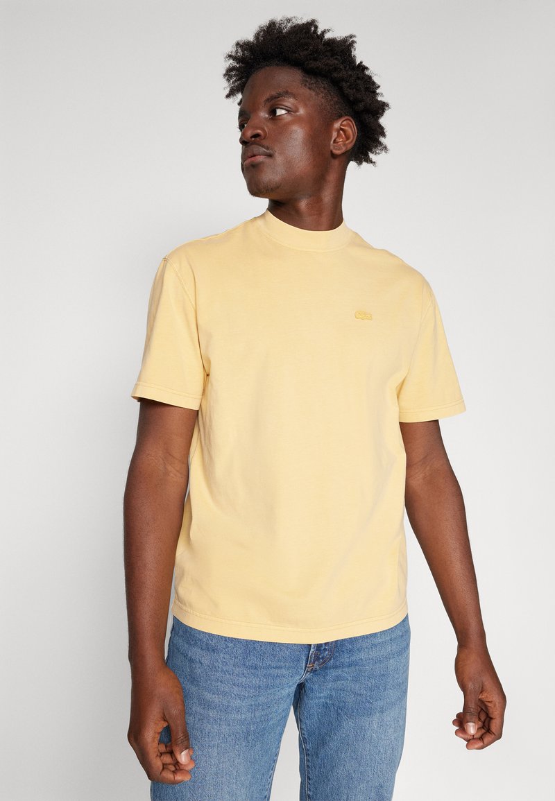 Jasnożółta bawełniana koszulka z okrągłym dekoltem i krótkimi rękawami. Ma na piersi małe, haftowane logo. Noszona z niebieskimi dżinsami.