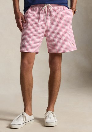 Mand iført røde og hvide ternede shorts med snøre, hvide sneakers og stående med hænderne i lommerne.