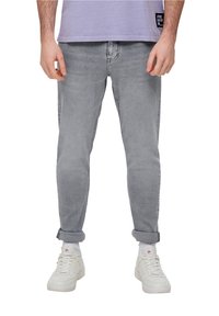 Jeans de mezclilla gris con un corte slim y puños enrollados, combinados con zapatillas blancas. La camisa es de color lila con una textura sutil y un detalle de logo.