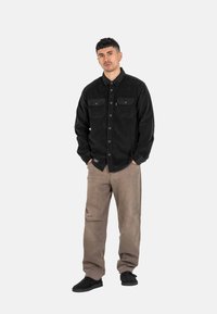 Schwarzes Cordhemd mit zwei Brusttaschen, Knopfverschluss und langen Ärmeln; kombiniert mit beigen, lockeren Hosen und schwarzen Schuhen.