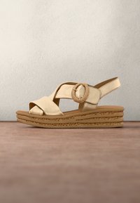 Sandale compensée beige avec semelle texturée, brides croisées, boucle ronde tressée et bride arrière sur une surface en bois.