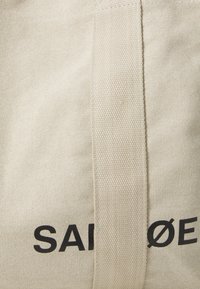 Borsa tote in tela leggermente beige, con testo stampato nero. Tessuto spesso e texture, con una maniglia piatta e robusta. Design minimalista.