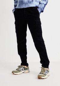 Czarne sztruksowe spodnie cargo o luźnym kroju z bocznymi kieszeniami, zestawione z wielokolorowymi trampkami w zielone i szare akcenty.