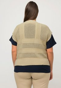 Beige gestrickte Weste mit einem Lochmuster, über einem dunkelblauen Oberteil mit kurzen Ärmeln getragen. Der untere Saum ist gerade und locker geschnitten.