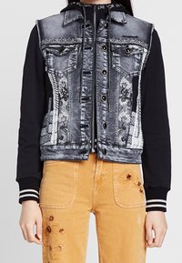 Jeansjacka med svarta ärmar, broderade mönster, två bröstfickor och silverknappar. Matchas med gula byxor med blommig broderi.