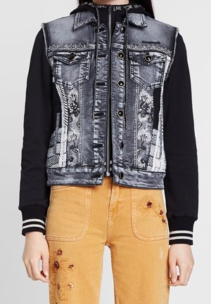 Giacca di jeans con maniche nere, modelli ricamati, due tasche sul petto e bottoni argentati. Abbinata a pantaloni gialli con ricami floreali.