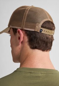 Gorra de béisbol de malla beige con visera curvada, correa ajustable en la parte trasera y un sutil detalle de marca. Lleva puesta una camisa verde oliva.