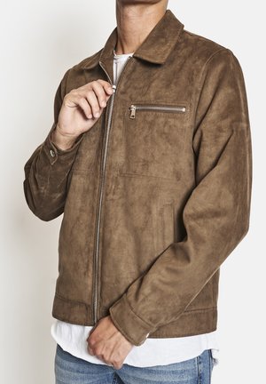 Mann trägt eine braune Wildlederjacke mit Frontreißverschluss und Brusttasche mit Reißverschluss, der Reißverschluss wird justiert, ein weißes Hemd und blaue Jeans sind sichtbar.