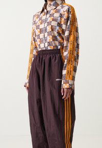 Långärmad skjorta i geometriskt mönstrad orange och brun tyg, parat med lösa bruna byxor med orangea sidostråk och elastisk midja.