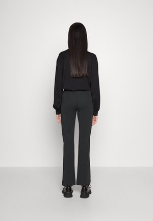 Vrouw met lang, stijl haar, gekleed in een zwart shirt met lange mouwen, donkere strakke broek en zwarte schoenen, staand tegen een effen witte achtergrond.