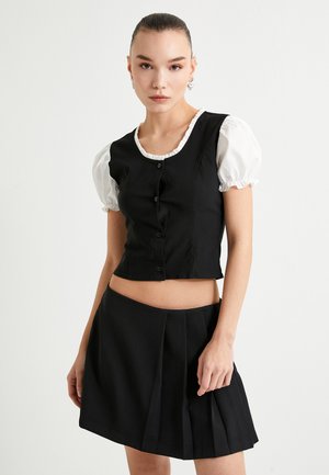 Schwarzer kurz geschnittener Weste mit Knöpfen und einem weißen Rüschenausschnitt, kombiniert mit einem schwarzen plissierten Minirock, präsentiert von einem Model.