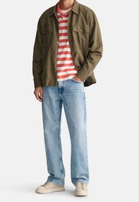 Olivgrön knäppjacka över en röd och vit randig polo, i kombination med ljusblå raka jeans och beige sneakers.