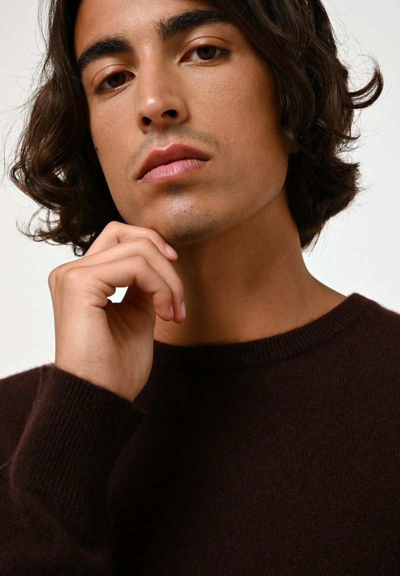 Brązowy sweter z okrągłym dekoltem, o fakturowanej tkaninie, zaprezentowany na osobie dotykającej podbródka z neutralnym wyrazem twarzy.