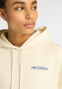 Un sweat à capuche crème fabriqué en tissu doux, arborant un logo bleu et rouge "new balance" sur la poitrine. Cordon de serrage à la capuche.