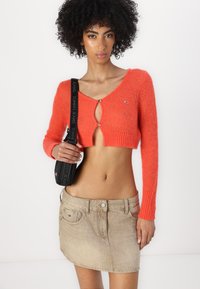 Bright orange cropped sweater with long sleeves and a keyhole front, paired with a light beige denim mini skirt and a black bag.