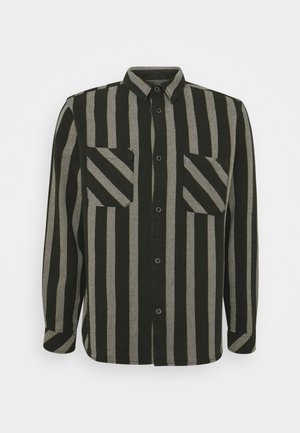 Chemise à manches longues boutonnée avec des rayures verticales noires et beiges et deux poches poitrine à rayures diagonales, sur un fond uni clair.