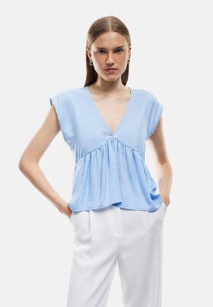 Mujer con cabello liso lleva una blusa sin mangas azul claro con escote en V y pantalones blancos, de pie con las manos en los bolsillos frente a fondo blanco.