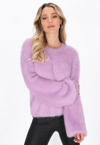 Donna con lunghi capelli biondi che indossa orecchini a cerchio, un maglione fluffy lavanda con piccole decorazioni e pantaloni neri, in posa con le braccia incrociate.