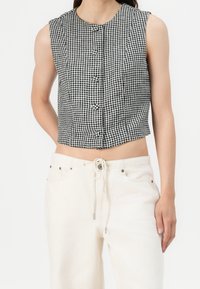 Ärmlös svart och vit rutigt croptop med frontknappar, tillsammans med vita byxor med dragsko och silverfärgade detaljer.