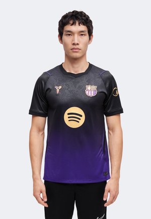 Jeune homme portant un maillot de sport noir et violet avec un logo Spotify doré et un écusson du club de football de Barcelone sur la poitrine.